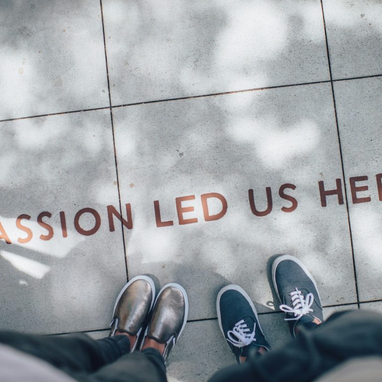 Vier Schritte – Passion Lead Us Here Vier Füße in Schuhen auf Asphalt mit dem Schriftzug "Passion lead us here".